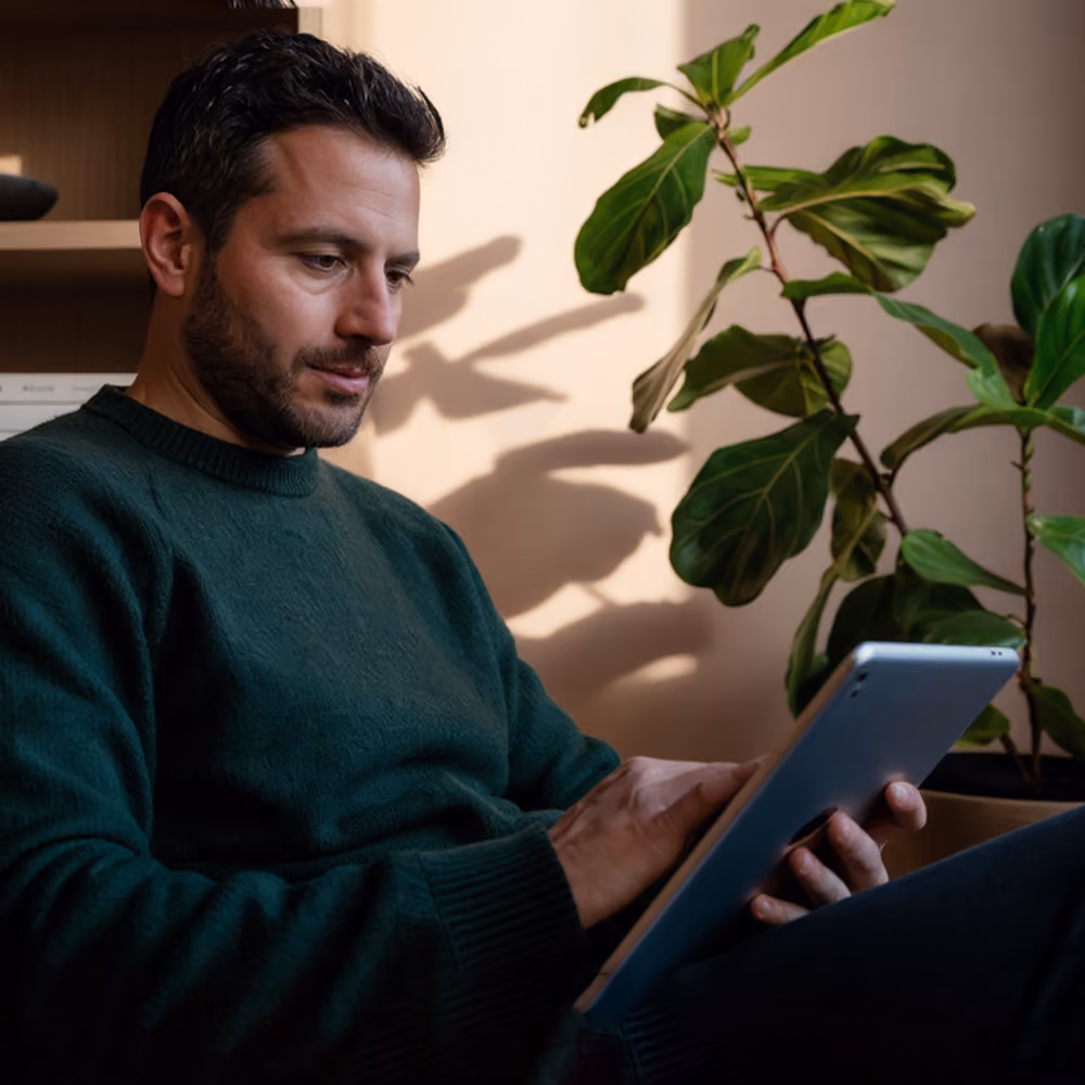 Man with dark hair and beard wearing a green sweater, sitting indoors while using a tablet, with a large leafy plant in the background.